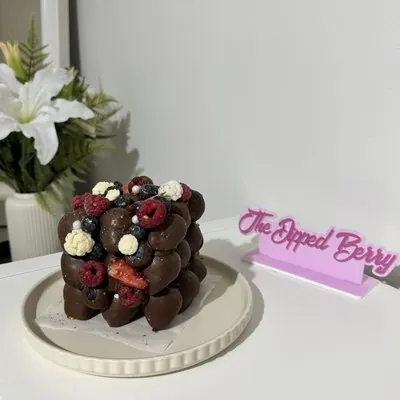 A stacked chocolate strawberry cake topped with fresh berries on a cream cake stand.